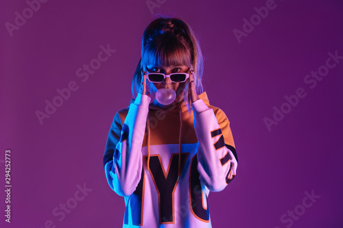 Pretty young 20s fashion teen girl model wear glasses blowing bubble gum looking at camera standing at purple studio background, igen teenager in trendy stylish night glow 80s 90s concept, portrait photo