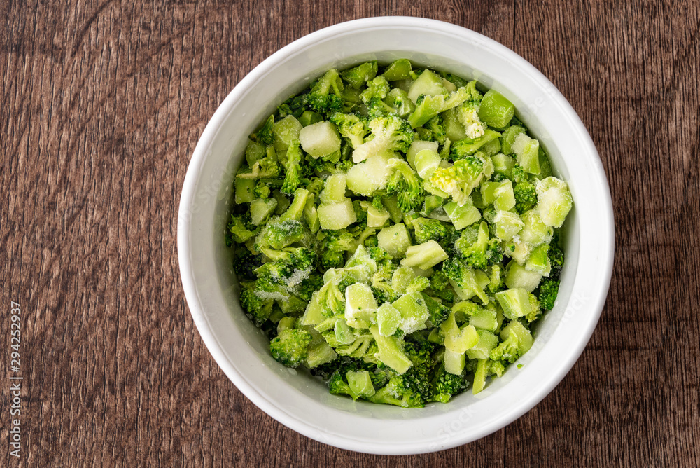 Round white bowl of chopped frozen broccoli on a dark wood background Round white bowl of chopped frozen broccoli on a dark wood background