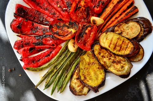 Grilled vegetables on a white plate. Appetizing grilled vegetables in a plate on a dark background, close-up. Vegetarian wholesome food. Baked eggplant, bell peppers, tomatoes, asparagus and carrots.