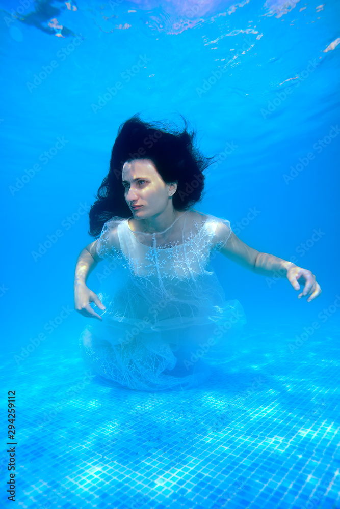 A beautiful girl in a white dress swims and poses underwater in the pool with her hair down on a Sunny day. Portrait. Unusual wedding. Underwater photography