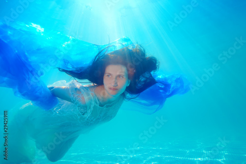 A surreal bride in a white dress swims and poses underwater in a pool with her hair down on a Sunny day with a blue cloth in her hands. Portrait. Unusual wedding. Underwater photography