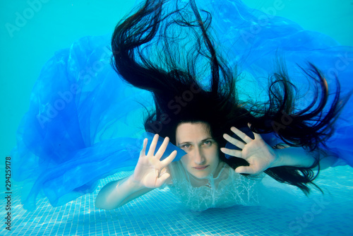 Portrait of an unusual bride who poses for the camera underwater at the bottom of the pool with her long hair down, in a white dress with a blue cloth in her hands. Portrait. Surrealism
