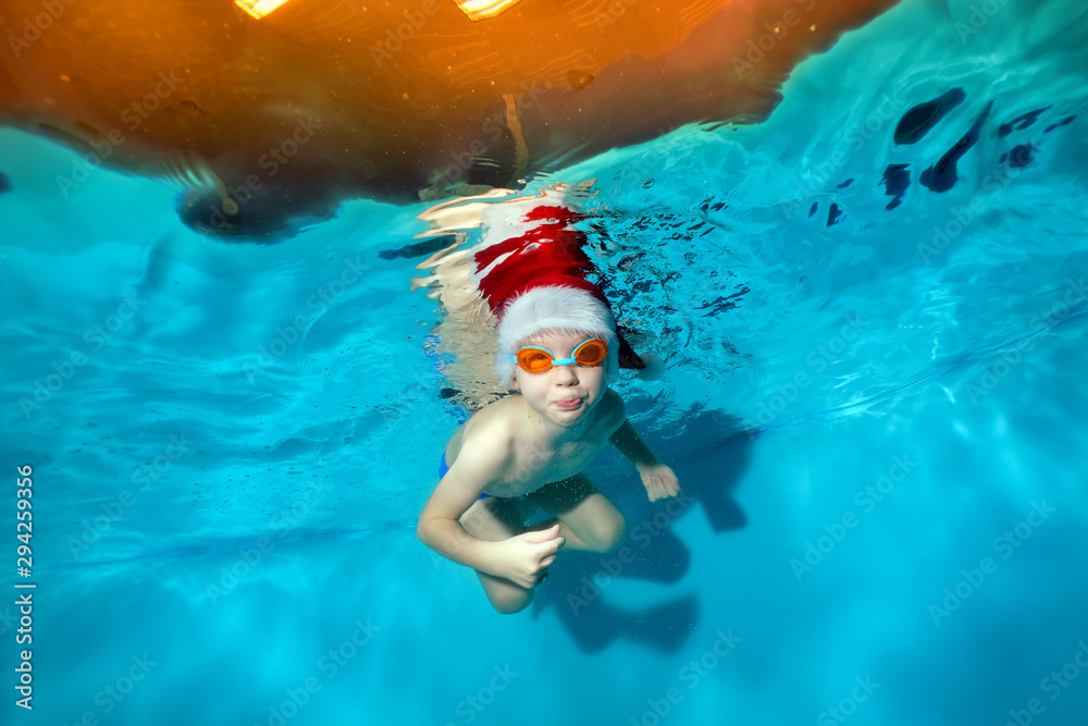 A smiling little boy poses underwater in a red Santa hat and swimming goggles. He looks into the camera and lets bubbles out on a blue background. Portrait. Concept. Underwater photography A smiling little boy poses underwater in a red Santa hat and swimming goggles. He looks into the camera and lets bubbles out on a blue background. Portrait. Concept. Underwater photography