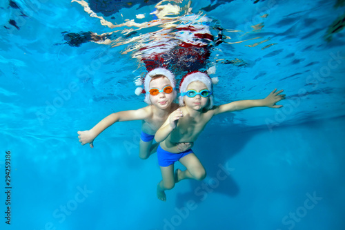 Two little boys swim and pose underwater in red Santa hats and swimming goggles. They hug, look at the camera and smile with their arms outstretched. Christmas celebration. Portrait. Concept