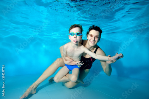 A little boy with his mother hugging and posing under the water at the bottom of the pool. They hug and look at the camera. A child with swimming glasses. Portrait. Family entertainment