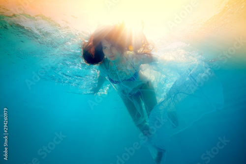 A beautiful girl swims underwater in the bright rays of the sun. She floats with her hair down against the bright yellow light from the surface. Digital art portrait. Bottom view