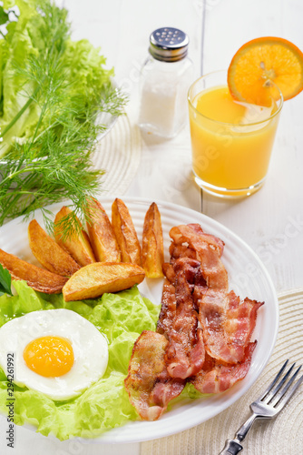 English breakfast-plate with fried bacon, with French fries, fried egg stands on a white table and a glass of fresh juice.