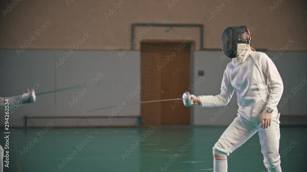 Two young women fencers in protective suits having a training in the gym in bright lights Two young women fencers in protective suits having a training in the gym in bright lights