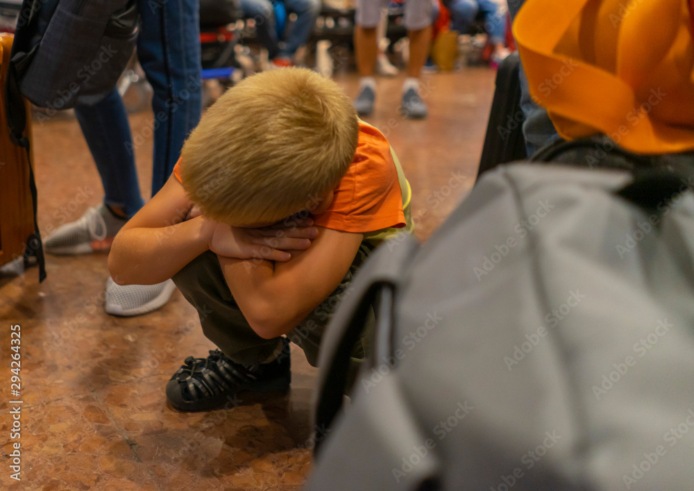 blonde boy tired of waiting for a delayed flight at the airport, crouching near baggage