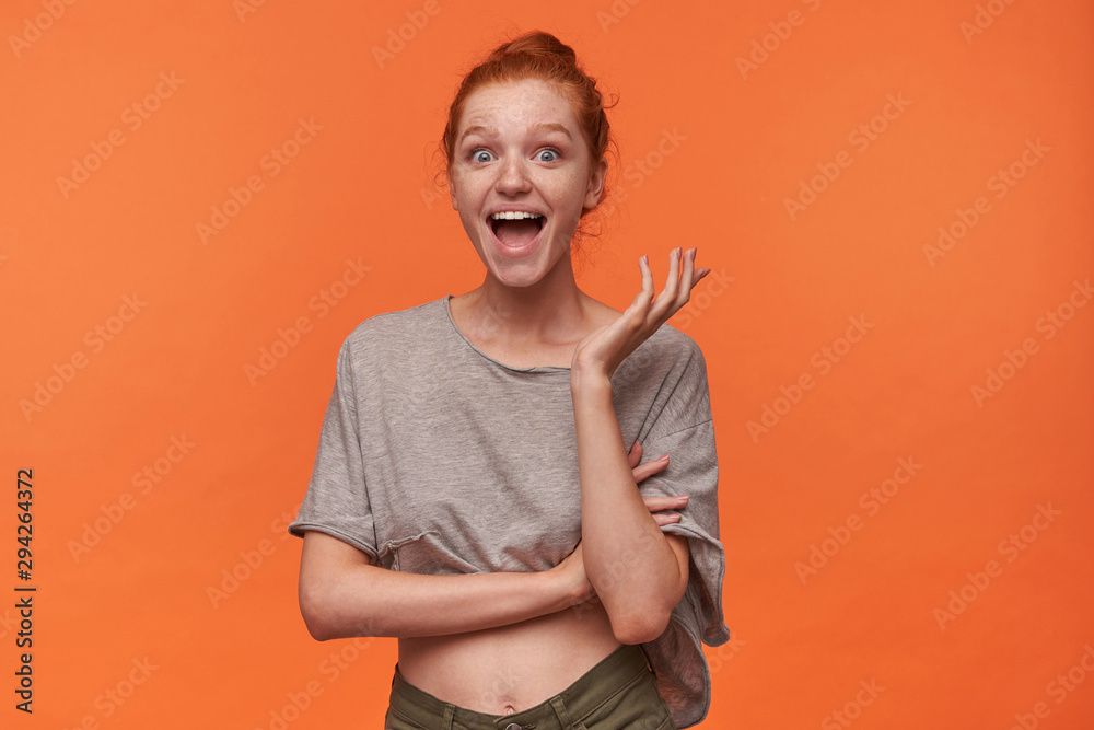 Studio photo of lovely joyful young readhead woman with bun hairstyle wearing grey t-shirt and green shorts, looking surprisedly to camera with wide mouth opened, standing over orange background - obrazy, fototapety, plakaty 