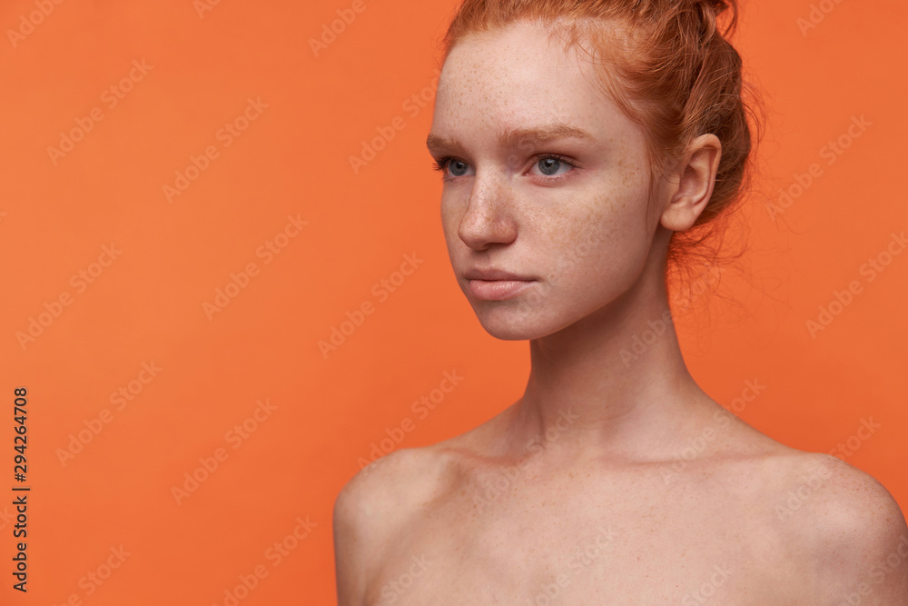 Close-up of charming young female wearing her foxy hair in knot looking aside with calm face, keeping her lips folded, posing over orange background with hands down Close-up of charming young female wearing her foxy hair in knot looking aside with calm face, keeping her lips folded, posing over orange background with hands down
