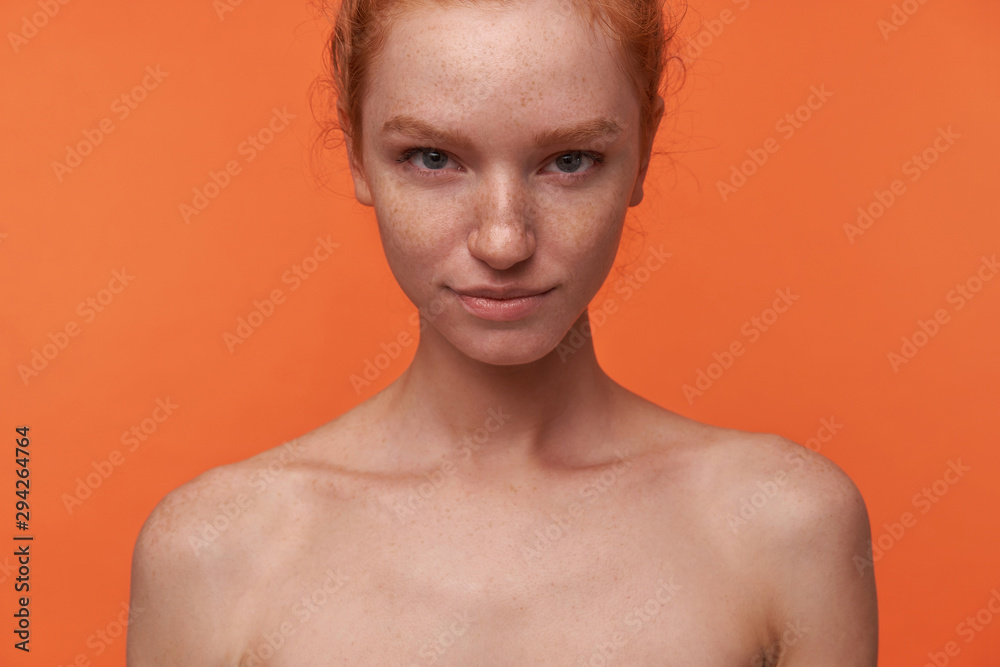Studio shot of beautiful young lady with red hair in knot standing over orange background with hands down, smiling softly to camera with raised eyebrow. Human emotion facial expression body language Studio shot of beautiful young lady with red hair in knot standing over orange background with hands down, smiling softly to camera with raised eyebrow. Human emotion facial expression body language