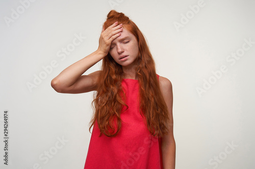 Portrait of attractive young readhead lady wearing with casual hairstyle standing over white background in pink dress, feeling sick and raising palm to her forehead, frowning with closed eyes