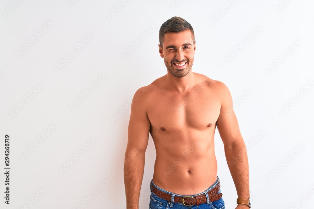Fototapeta premium Young handsome shirtless man showing muscular body over isolated background winking looking at the camera with sexy expression, cheerful and happy face.