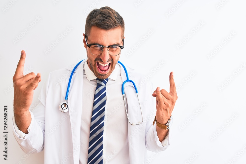 Young handsome doctor man wearing stethoscope over isolated background shouting with crazy expression doing rock symbol with hands up. Music star. Heavy concept.