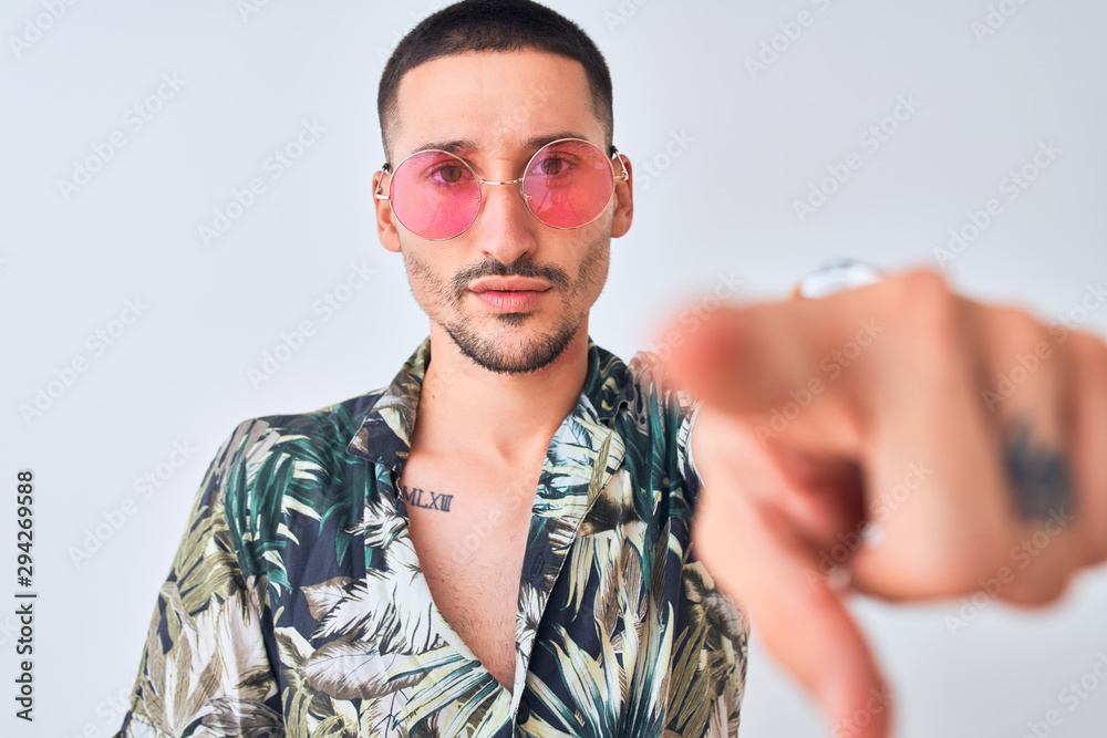 Young handsome man wearing Hawaiian sumer shirt and pink sunglasses over isolated background pointing with finger to the camera and to you, hand sign, positive and confident gesture from the front Young handsome man wearing Hawaiian sumer shirt and pink sunglasses over isolated background pointing with finger to the camera and to you, hand sign, positive and confident gesture from the front