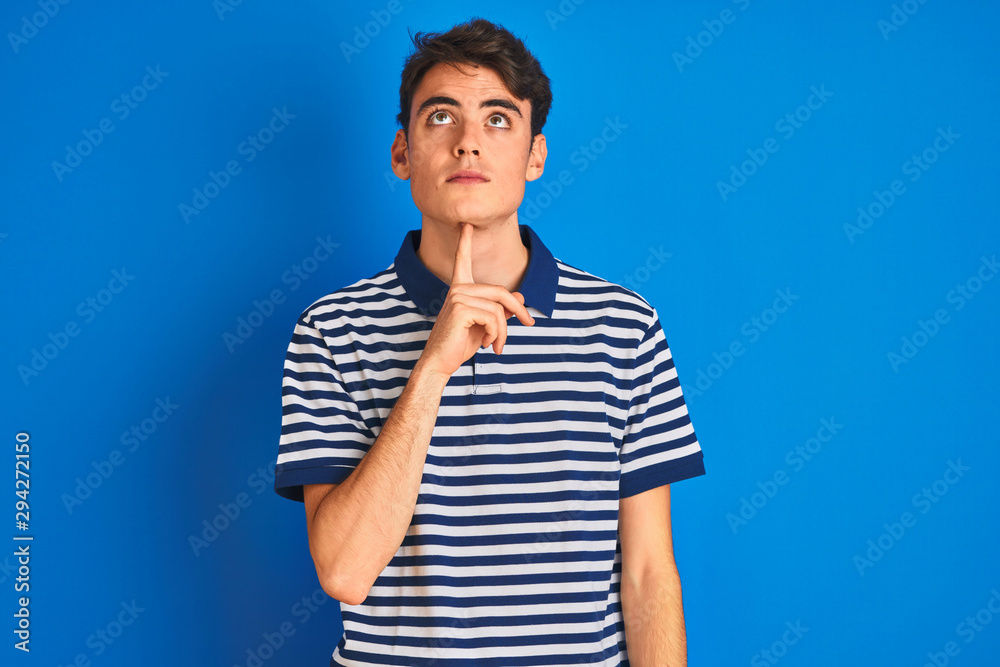 Teenager boy wearing casual t-shirt standing over blue isolated background Thinking concentrated about doubt with finger on chin and looking up wondering Teenager boy wearing casual t-shirt standing over blue isolated background Thinking concentrated about doubt with finger on chin and looking up wondering