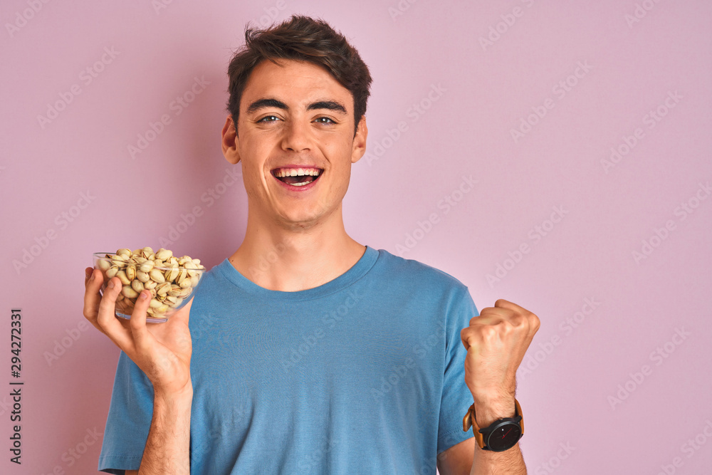 Teenager boy holding a bunch of pistachios over isolated pink background screaming proud and celebrating victory and success very excited, cheering emotion Teenager boy holding a bunch of pistachios over isolated pink background screaming proud and celebrating victory and success very excited, cheering emotion