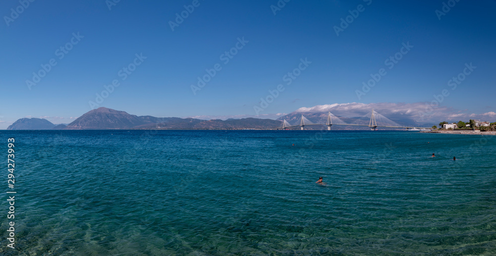 Bright blue ocean with mountains and a bridge in the background and people swimming in the water.