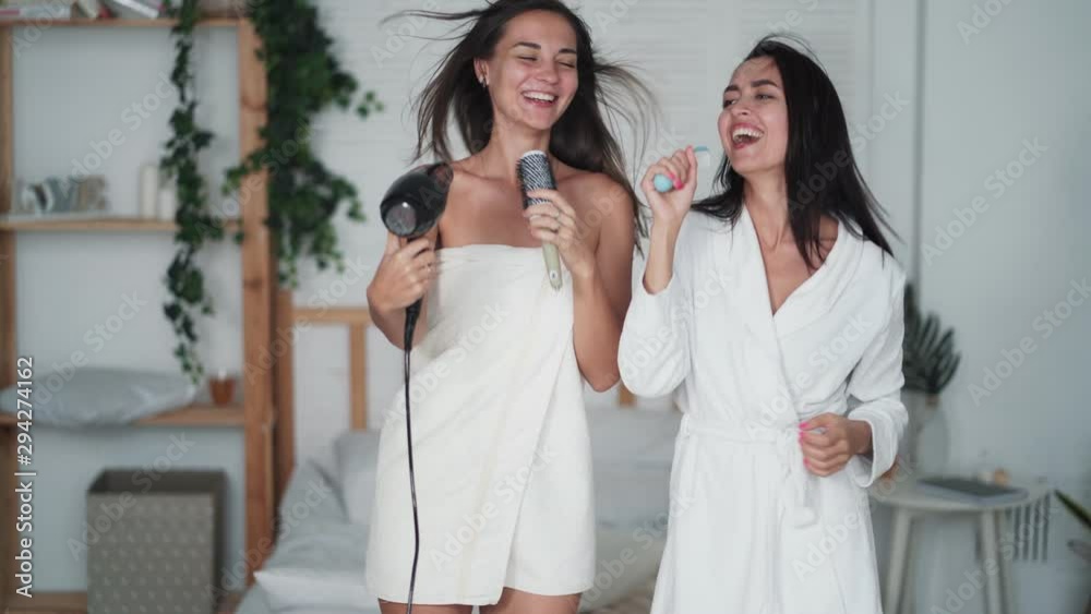 Two girl friends in white bathrobe dance, sing and dry hair with hairdryer, laugh Two girl friends in white bathrobe dance, sing and dry hair with hairdryer, laugh