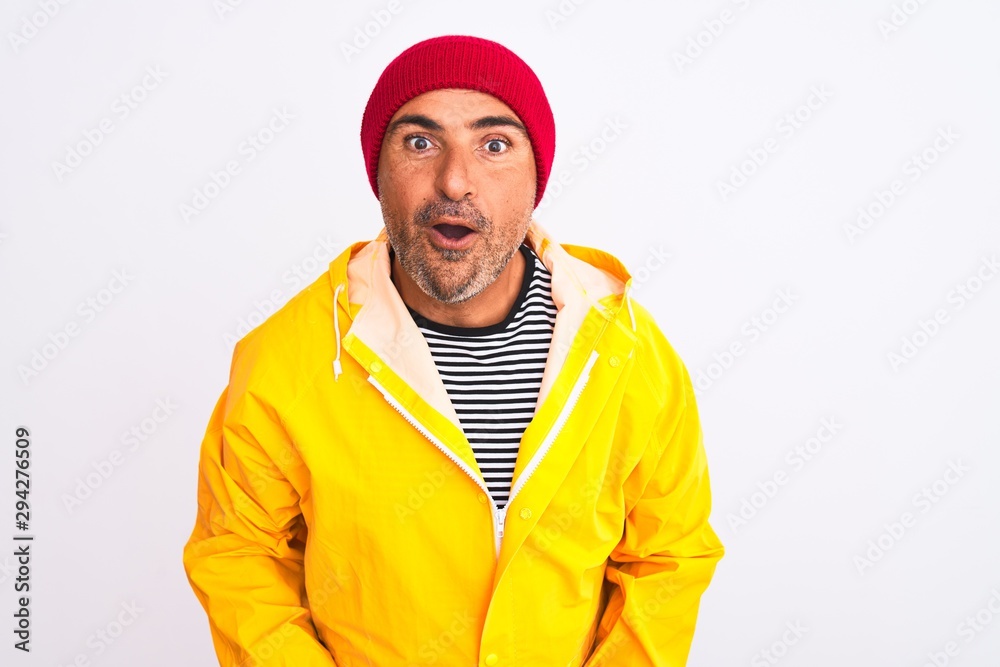 Middle age man wearing rain coat and woolen hat standing over isolated white background afraid and shocked with surprise expression, fear and excited face.