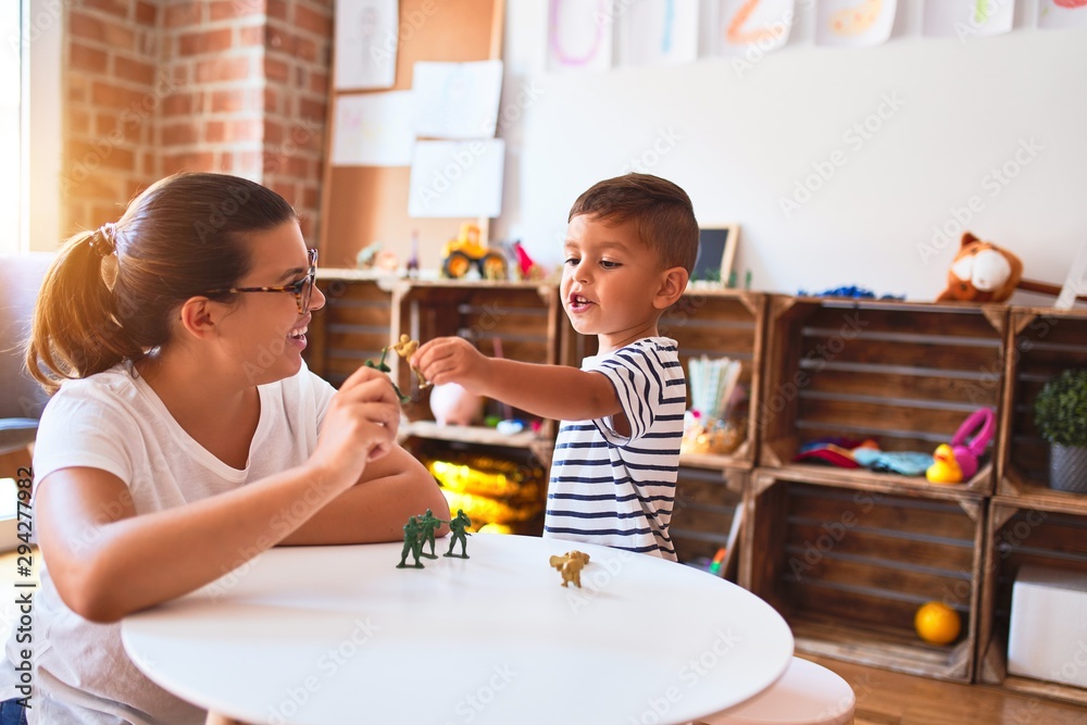 Beautiful teacher and toddler boy playing with figurine army soldiers at kindergarten - obrazy, fototapety, plakaty 