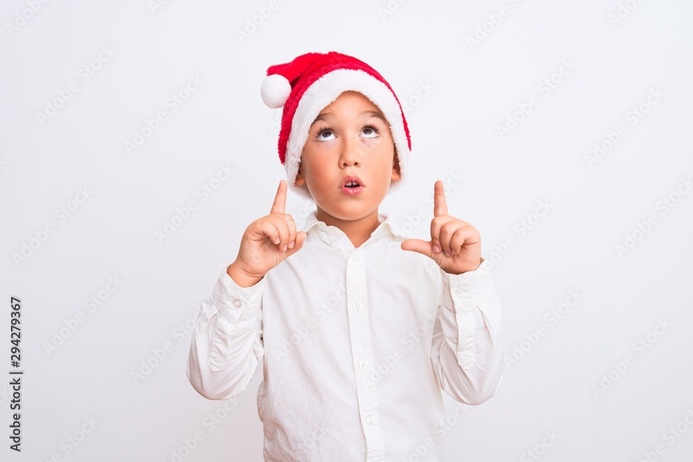 Beautiful kid boy wearing Christmas Santa hat standing over isolated white background amazed and surprised looking up and pointing with fingers and raised arms. Beautiful kid boy wearing Christmas Santa hat standing over isolated white background amazed and surprised looking up and pointing with fingers and raised arms.
