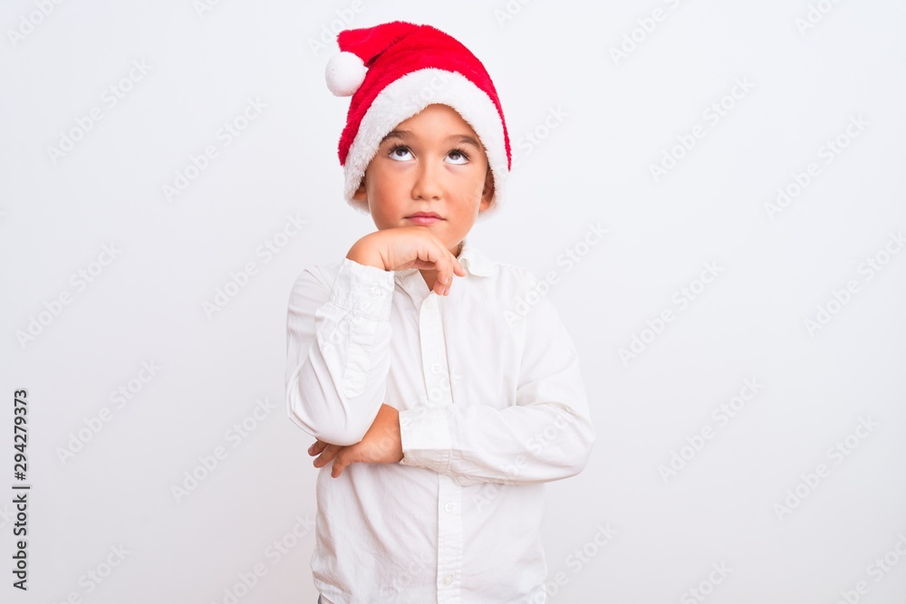 Beautiful kid boy wearing Christmas Santa hat standing over isolated white background with hand on chin thinking about question, pensive expression. Smiling with thoughtful face. Doubt concept.