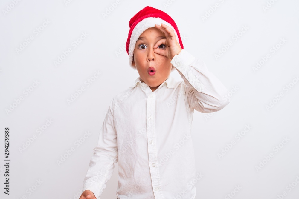 Beautiful kid boy wearing Christmas Santa hat standing over isolated white background doing ok gesture shocked with surprised face, eye looking through fingers. Unbelieving expression. Beautiful kid boy wearing Christmas Santa hat standing over isolated white background doing ok gesture shocked with surprised face, eye looking through fingers. Unbelieving expression.