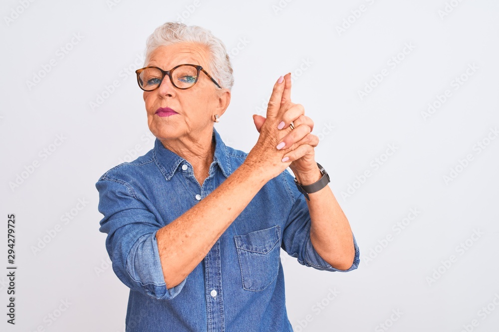 Senior grey-haired woman wearing denim shirt and glasses over isolated white background Holding symbolic gun with hand gesture, playing killing shooting weapons, angry face