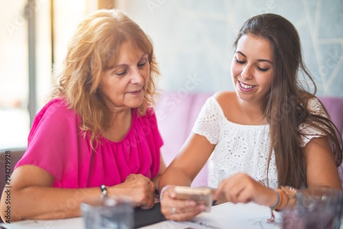 Beautiful mother and daugther sitting at restaurant using smartphone smiling