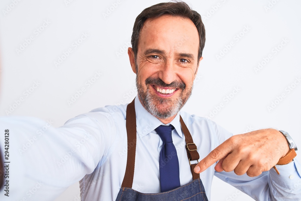 Middle age shopkeeper man wearing apron make selfie over isolated white background very happy pointing with hand and finger Middle age shopkeeper man wearing apron make selfie over isolated white background very happy pointing with hand and finger