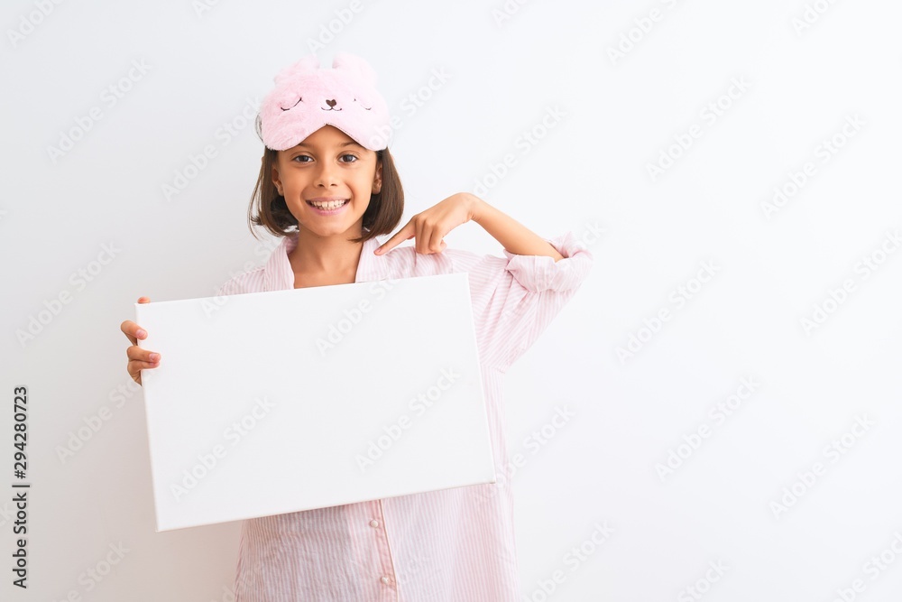 Child girl wearing sleep mask and pajama holding banner over isolated white background with surprise face pointing finger to himself Child girl wearing sleep mask and pajama holding banner over isolated white background with surprise face pointing finger to himself