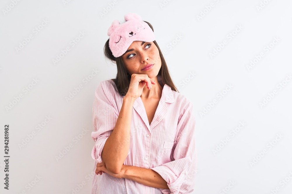 Young beautiful woman wearing sleep mask and pajama over isolated white background with hand on chin thinking about question, pensive expression. Smiling with thoughtful face. Doubt concept. Young beautiful woman wearing sleep mask and pajama over isolated white background with hand on chin thinking about question, pensive expression. Smiling with thoughtful face. Doubt concept.