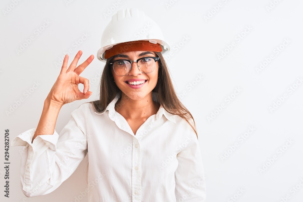 Young beautiful architect woman wearing helmet and glasses over isolated white background smiling positive doing ok sign with hand and fingers. Successful expression. Young beautiful architect woman wearing helmet and glasses over isolated white background smiling positive doing ok sign with hand and fingers. Successful expression.