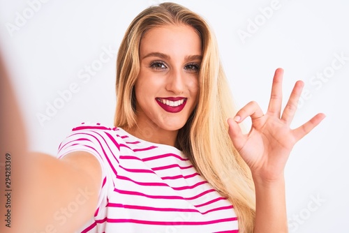 Beautiful woman wearing striped t-shirt make selfie by camera over isolated white background doing ok sign with fingers, excellent symbol