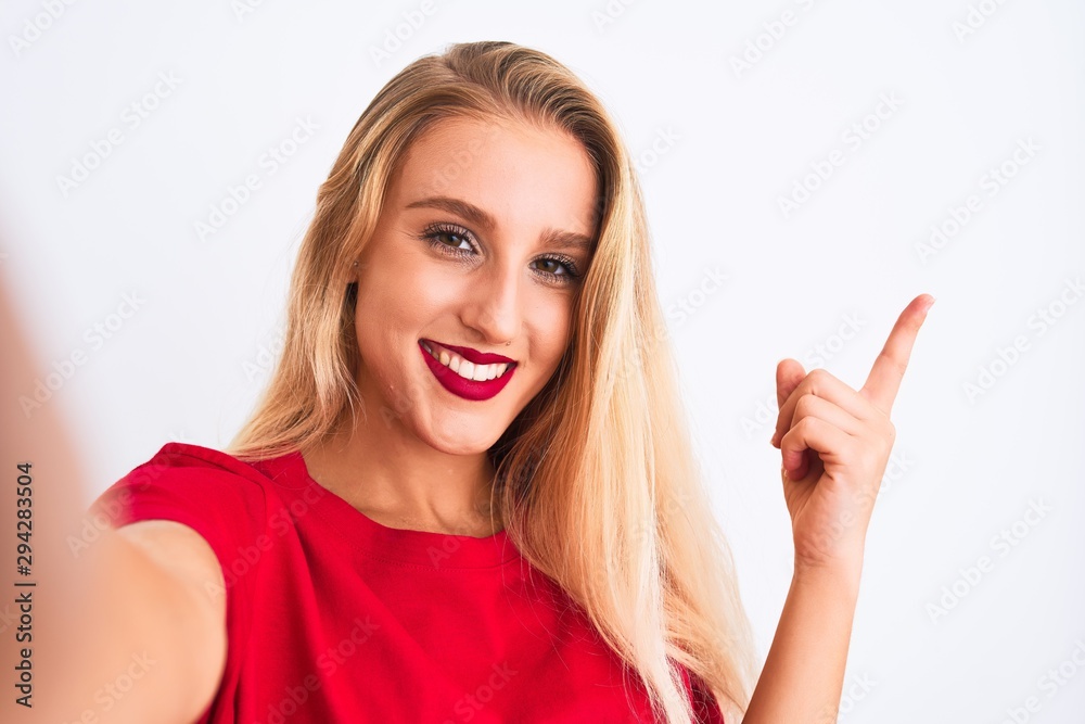 Young beautiful woman wearing t-shirt make selfie by camera over isolated white background very happy pointing with hand and finger to the side Young beautiful woman wearing t-shirt make selfie by camera over isolated white background very happy pointing with hand and finger to the side