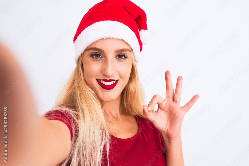 Woman wearing Christmas Santa hat make selfie by camera over isolated white background doing ok sign with fingers, excellent symbol Woman wearing Christmas Santa hat make selfie by camera over isolated white background doing ok sign with fingers, excellent symbol