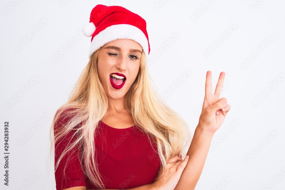 Young beautiful woman wearing Christmas Santa hat over isolated white background smiling with happy face winking at the camera doing victory sign. Number two. Young beautiful woman wearing Christmas Santa hat over isolated white background smiling with happy face winking at the camera doing victory sign. Number two.