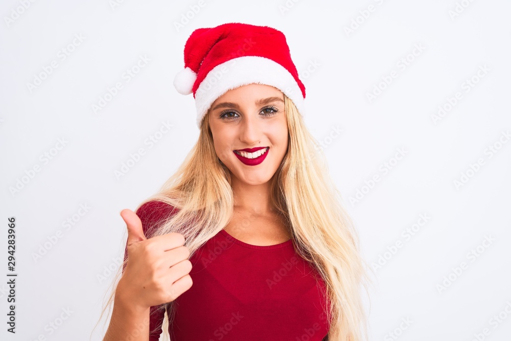 Young beautiful woman wearing Christmas Santa hat over isolated white background doing happy thumbs up gesture with hand. Approving expression looking at the camera showing success. Young beautiful woman wearing Christmas Santa hat over isolated white background doing happy thumbs up gesture with hand. Approving expression looking at the camera showing success.