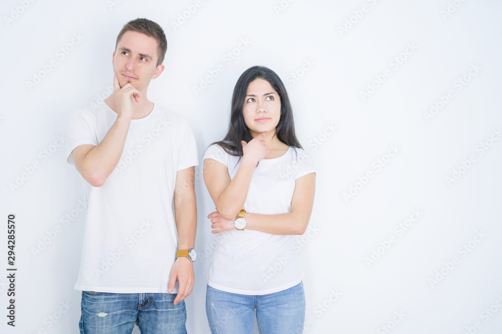 Young beautiful couple wearing casual t-shirt standing over isolated white background with hand on chin thinking about question, pensive expression. Smiling with thoughtful face. Doubt concept. Young beautiful couple wearing casual t-shirt standing over isolated white background with hand on chin thinking about question, pensive expression. Smiling with thoughtful face. Doubt concept.