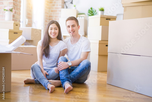 Young beautiful couple sitting on the floor at new home around cardboard boxes with a happy face standing and smiling with a confident smile showing teeth