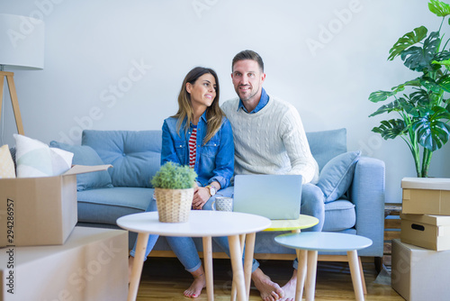 Young beautiful couple sitting on the sofa drinking cup of coffee using laptop at new home around cardboard boxes