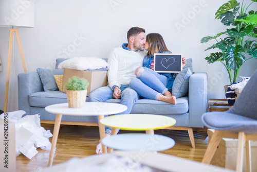 Young beautiful couple sitting on the sofa holding blackboard with a heart and kissing at new home around cardboard boxes