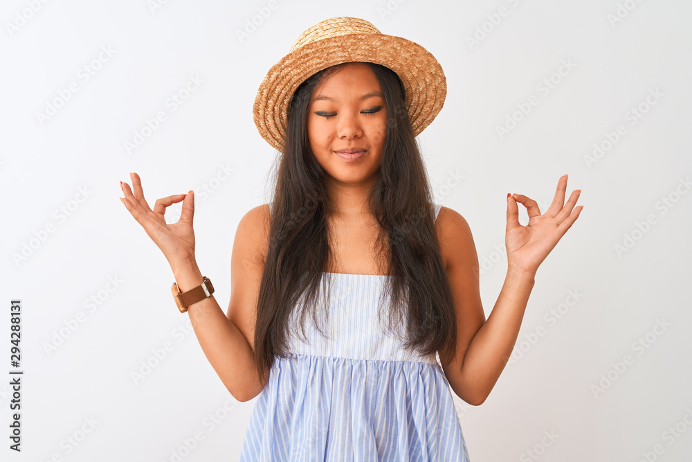 Young chinese woman wearing striped dress and hat standing over isolated white background relax and smiling with eyes closed doing meditation gesture with fingers. Yoga concept. Young chinese woman wearing striped dress and hat standing over isolated white background relax and smiling with eyes closed doing meditation gesture with fingers. Yoga concept.