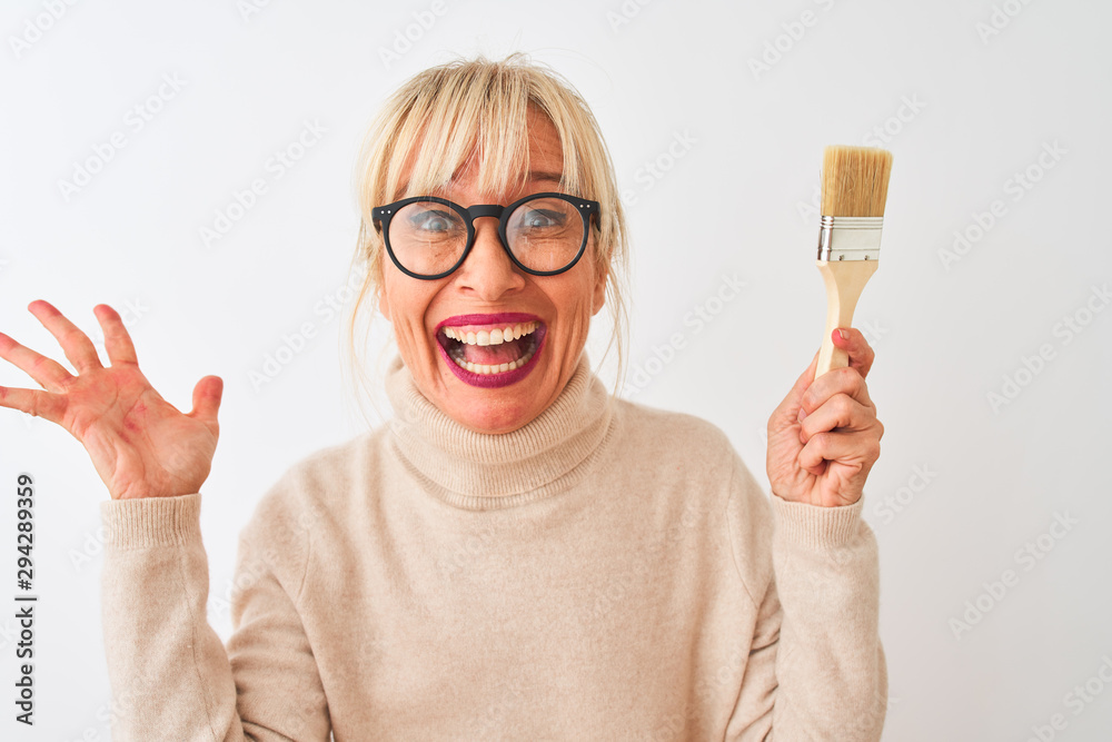 Middle age painter woman holding paint brush standing over isolated white background very happy and excited, winner expression celebrating victory screaming with big smile and raised hands Middle age painter woman holding paint brush standing over isolated white background very happy and excited, winner expression celebrating victory screaming with big smile and raised hands
