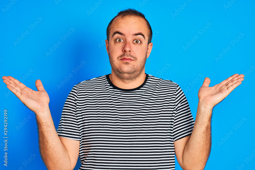 Young man wearing striped navy t-shirt standing over isolated blue background clueless and confused expression with arms and hands raised. Doubt concept. Young man wearing striped navy t-shirt standing over isolated blue background clueless and confused expression with arms and hands raised. Doubt concept.