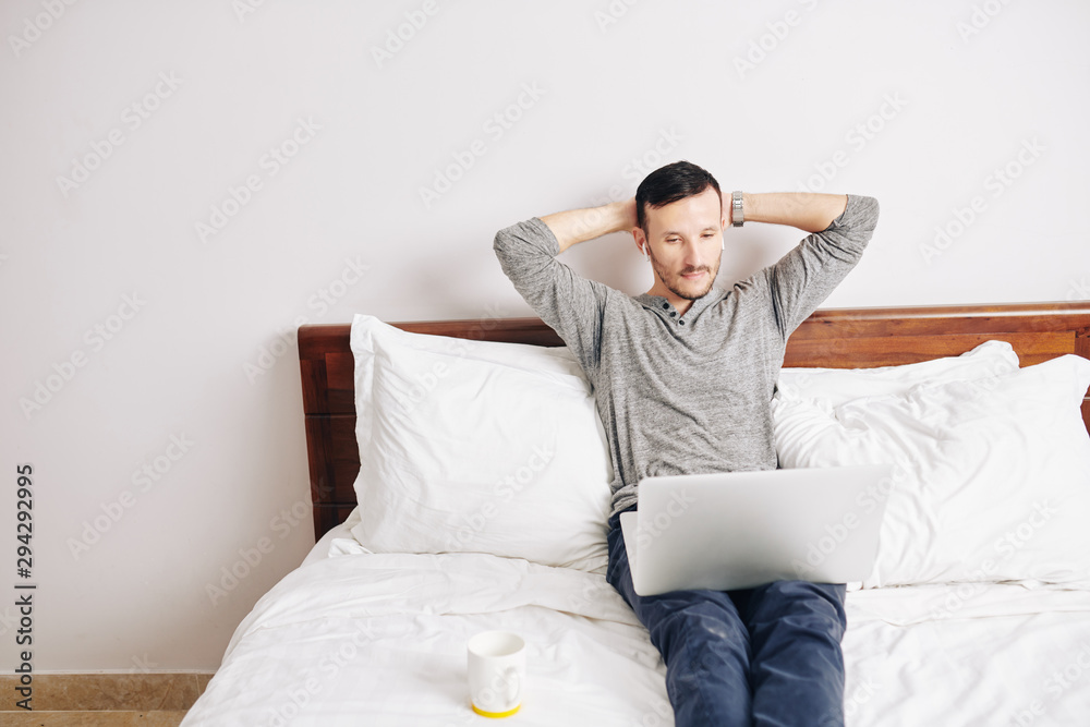 Handsome bearded man leaning back in his bed when watching new episode of popular show online