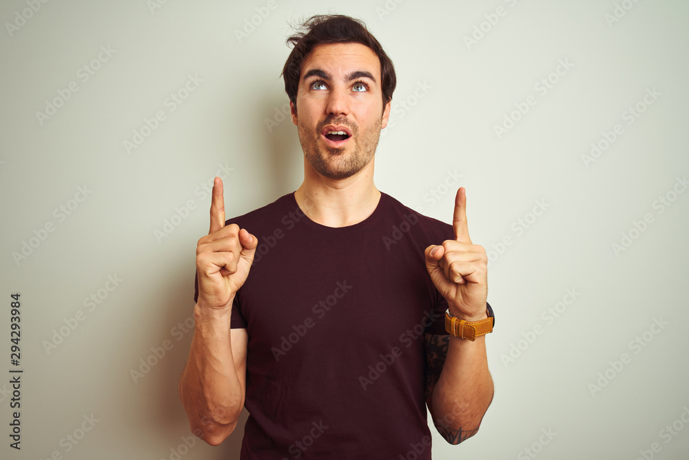 Young handsome man with tattoo wearing purple casual t-shirt over isolated white background amazed and surprised looking up and pointing with fingers and raised arms. Young handsome man with tattoo wearing purple casual t-shirt over isolated white background amazed and surprised looking up and pointing with fingers and raised arms.