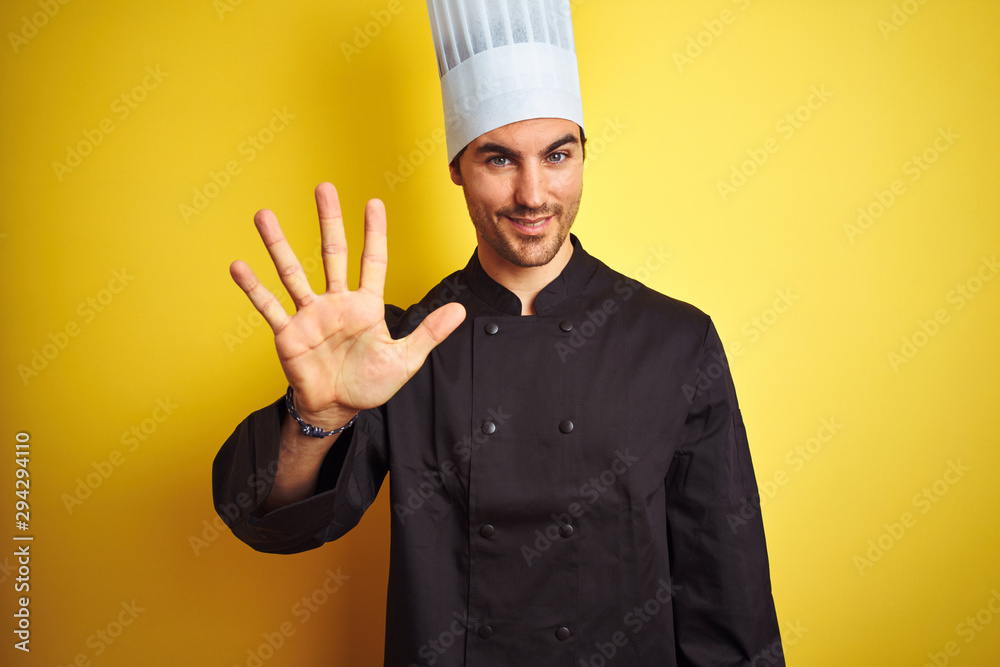 Young chef man wearing uniform and hat standing over isolated yellow background showing and pointing up with fingers number five while smiling confident and happy.